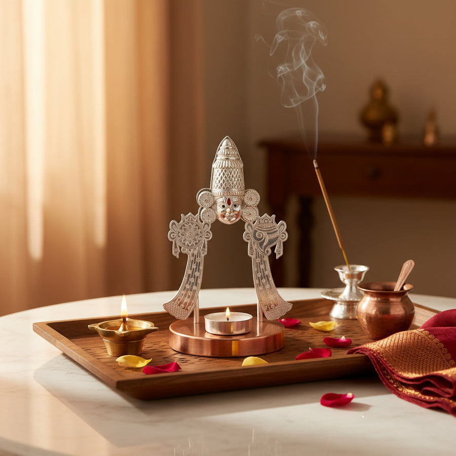 A decorative silver Shiva Lingam on a copper-colored base, styled on a marble surface with an incense holder, incense stick, copper pot, spoon, and red flower petals in a warm indoor setting.