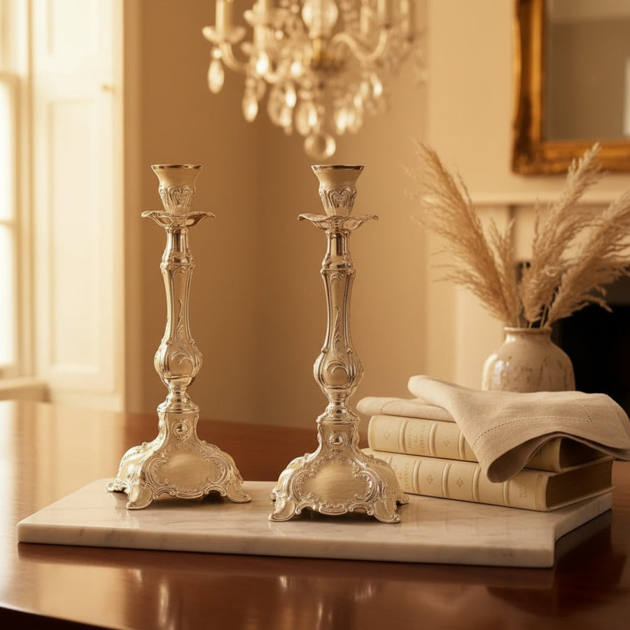 A pair of ornate silver candlesticks displayed on a polished wooden table in a dining room setting, with a wine glass, books, and a folded napkin nearby.