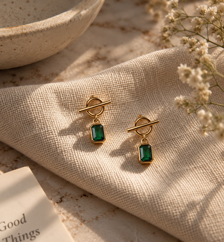 A pair of gold-toned drop earrings with rectangular green gemstones, styled on a textured beige fabric with dried flowers and a ceramic bowl in the background.