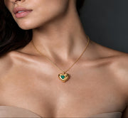 A close-up of a woman wearing a delicate gold heart pendant necklace with a green heart in the center, set against a dark gray background.