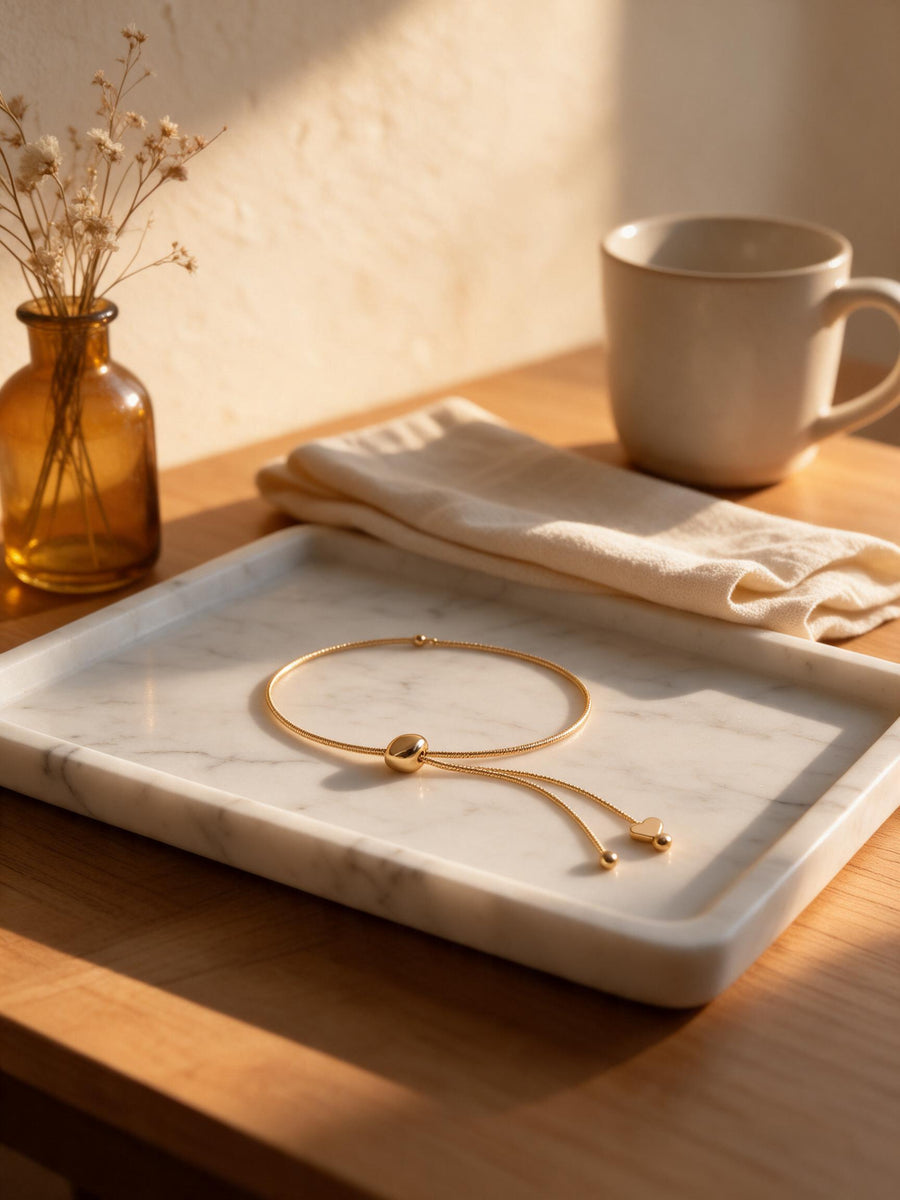 A delicate gold bangle bracelet with a central bead and sliding knot closure, displayed on a white marble tray on a wooden table. The scene is styled with a beige ceramic mug, a folded cream linen napkin, and a small amber glass vase with dried flowers, all illuminated by warm, directional sunlight.
