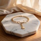 A gold chain bracelet with three dangling star charms, displayed on a white marble hexagonal tray over a wooden surface with soft cream fabric in the background.