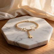 A gold chain bracelet with three dangling star charms, displayed on a white marble hexagonal tray over a wooden surface with soft cream fabric in the background.