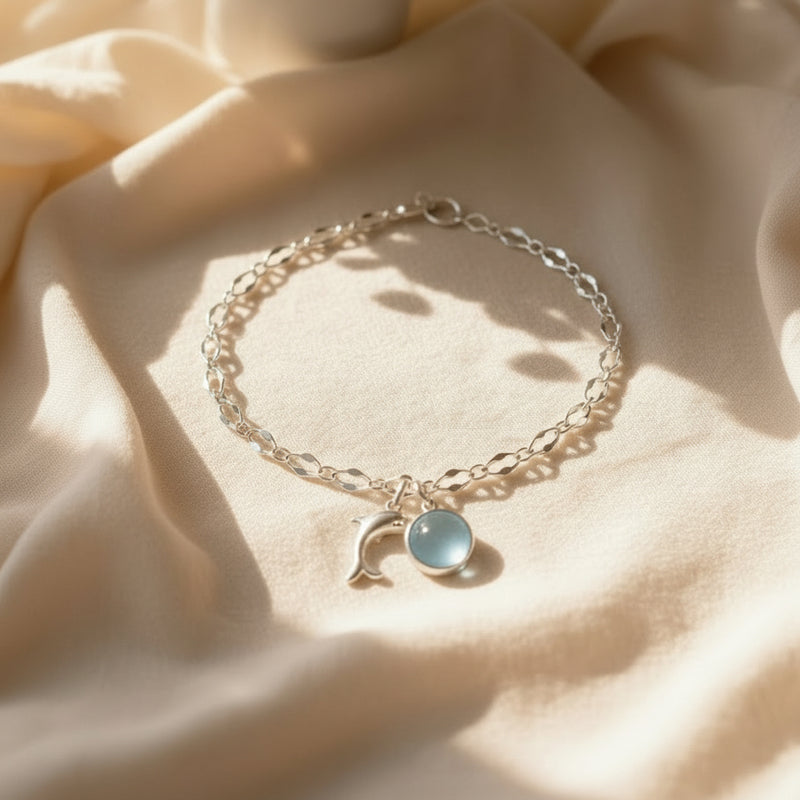 A silver-toned beaded bracelet with a light blue teardrop pendant rests on a cream ceramic plate, styled on a marble dresser with a flower arrangement, a mirror, and soft sunlight.