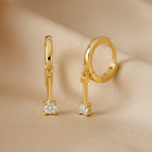 A pair of gold huggie hoop earrings with a dangling key-shaped charm, each featuring a clear round-cut gemstone, shown on a plain white background.