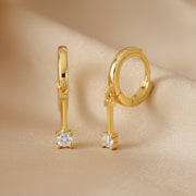 A pair of gold huggie hoop earrings with a dangling key-shaped charm, each featuring a clear round-cut gemstone, shown on a plain white background.