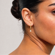 A close-up of a woman with dark hair in a bun wearing a delicate gold dangle earring with a small clear stone, set against a neutral light gray background.