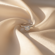 A close-up of a woman's hand wearing a delicate silver ring with a small clear gemstone, set against a soft beige background.