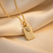 A gold rectangular pendant necklace with a small star detail and a clear central gemstone, resting on draped beige satin fabric.