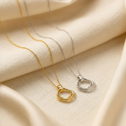 Two minimalist pendant necklaces draped on cream-colored fabric, one in polished gold and one in polished silver, both featuring an open, multi-layered geometric design.