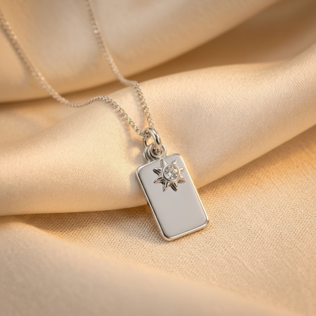 A silver rectangular pendant necklace with a small sunburst charm and clear gemstone, resting on draped beige satin fabric.