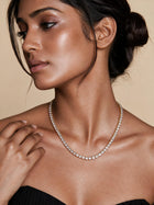 A close-up of a woman wearing a delicate diamond tennis necklace with a silver-toned setting, styled with a black strapless top against a warm beige background.