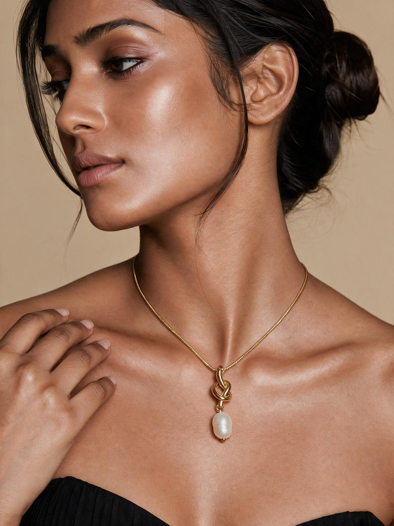 A close-up of a woman wearing a delicate gold chain necklace with a circular pendant featuring a black and white zebra-like pattern, set against a warm beige background.