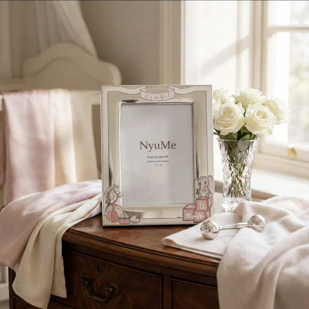 A silver picture frame with decorative cartoon bear illustrations and a 'It's a girl' banner, styled on a wooden chest next to white roses in a vase.