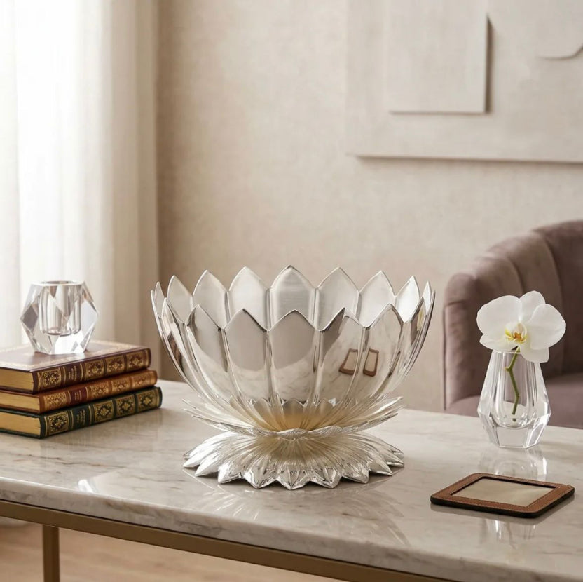 A lotus-shaped clear glass bowl with a flared pedestal base, styled on a marble coffee table in a living room setting alongside books, a small glass candle holder, and a vase with a white orchid.