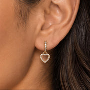 A close-up of a model's ear wearing a delicate gold huggie hoop earring with a dangling open heart charm set with small clear stones.