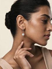 A close-up side profile of a woman wearing a delicate silver dangle earring with a small white heart-shaped pearl, set against a soft beige background.