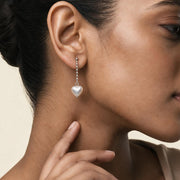 A close-up side profile of a woman wearing a delicate silver dangle earring with a small white heart-shaped pearl, set against a soft beige background.