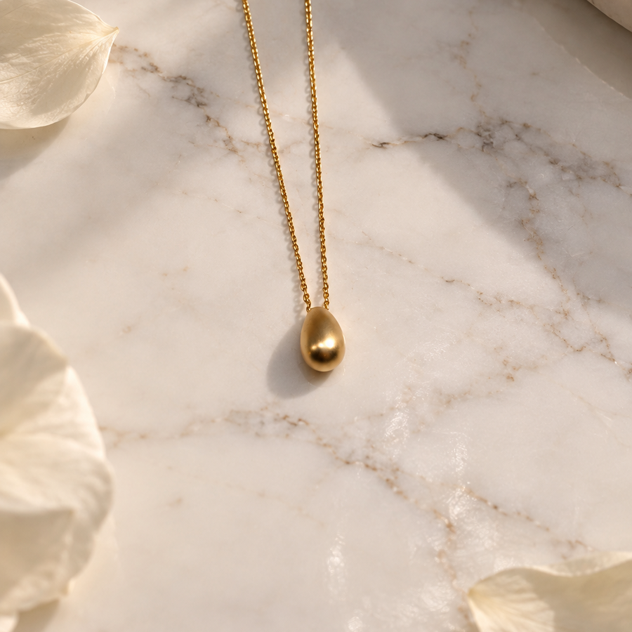 A delicate gold chain necklace with a small teardrop-shaped gold pendant, displayed on a white marble surface with white flower petals.