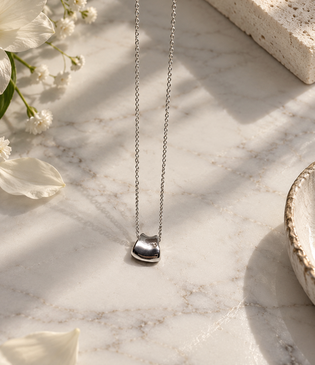 A silver heart pendant necklace on a delicate silver chain, displayed on a white marble surface with soft shadows, white flowers, and a textured stone prop.