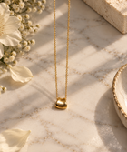 A delicate gold chain necklace with a small, polished gold bead pendant, styled on a light marble surface with white flowers and a textured stone block.
