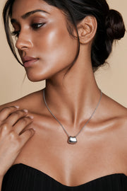 A close-up lifestyle shot of a model wearing a delicate gold chain necklace with a small, polished gold pendant against a warm beige background.