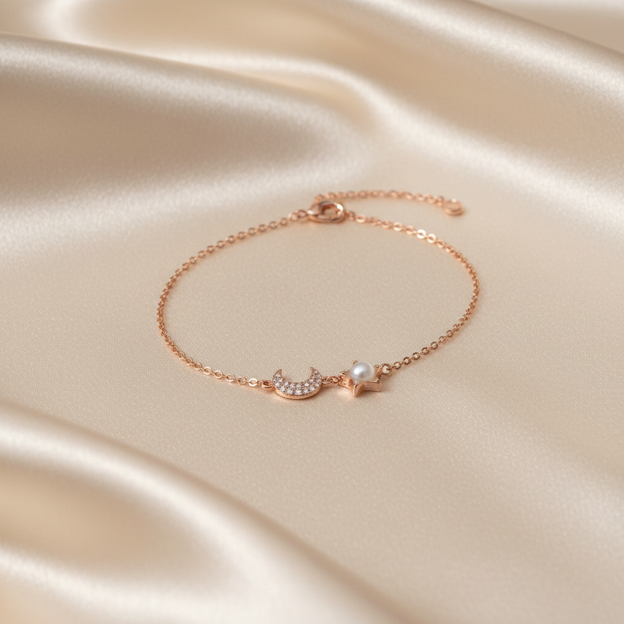 A delicate rose gold chain bracelet featuring a crescent moon and star charm, one of which is set with a small white pearl, displayed on a draped champagne satin fabric.