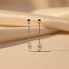 A pair of silver dangle earrings with heart-shaped charms, displayed on a white textured foam block held by a hand.