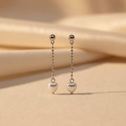 A pair of silver dangle earrings with heart-shaped charms, displayed on a white textured foam block held by a hand.