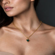 A close-up of a woman wearing a delicate gold chain necklace with a small heart-shaped pendant featuring a dark green inlay, set against a dark gray background.