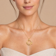 A close-up of a woman wearing a delicate gold chain necklace with a double interlocking ring pendant, set against a warm beige background.